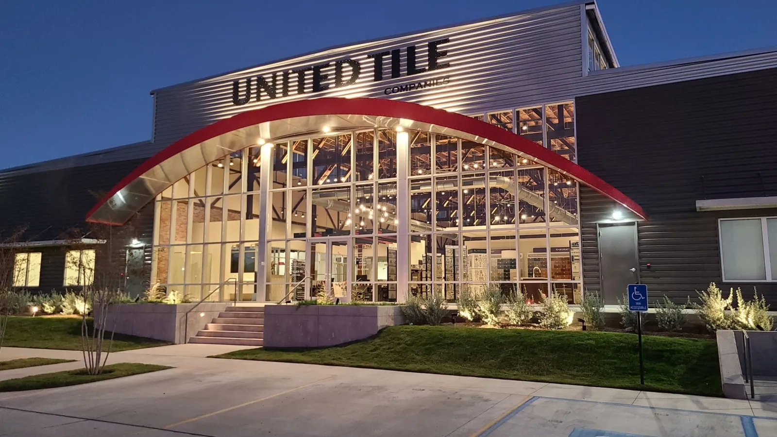 Our home today: the United Tile building at dusk, its red arch and two-story wall of glass lit from inside, with the showroom and timber trusses visible through the windows.