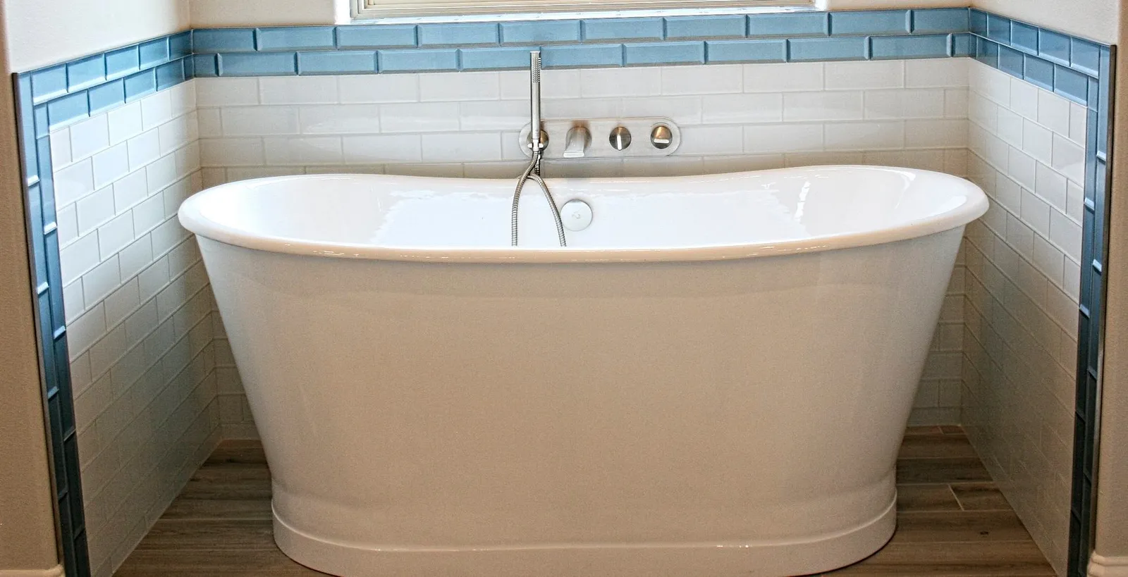 Freestanding tub against white tile with a blue subway tile accent band.