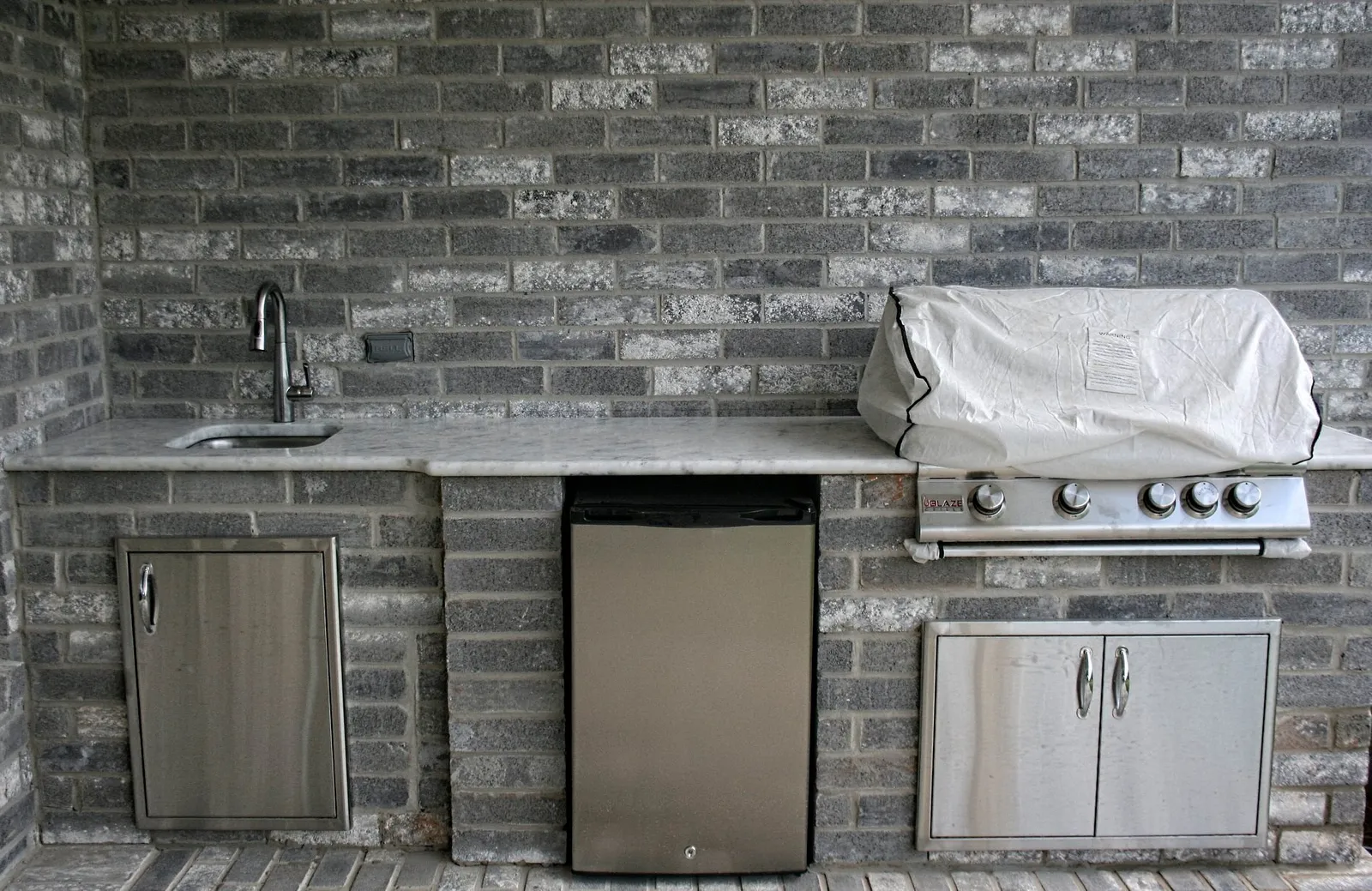Outdoor kitchen with a gray brick-look tile wall, built-in grill and fridge.