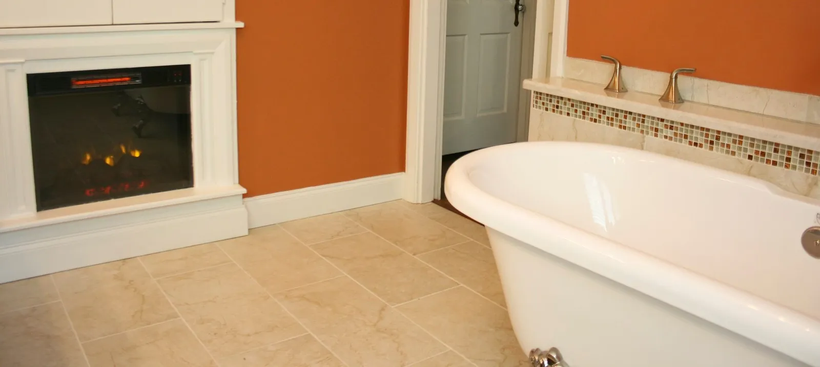 Clawfoot tub beside an electric fireplace on beige tile.