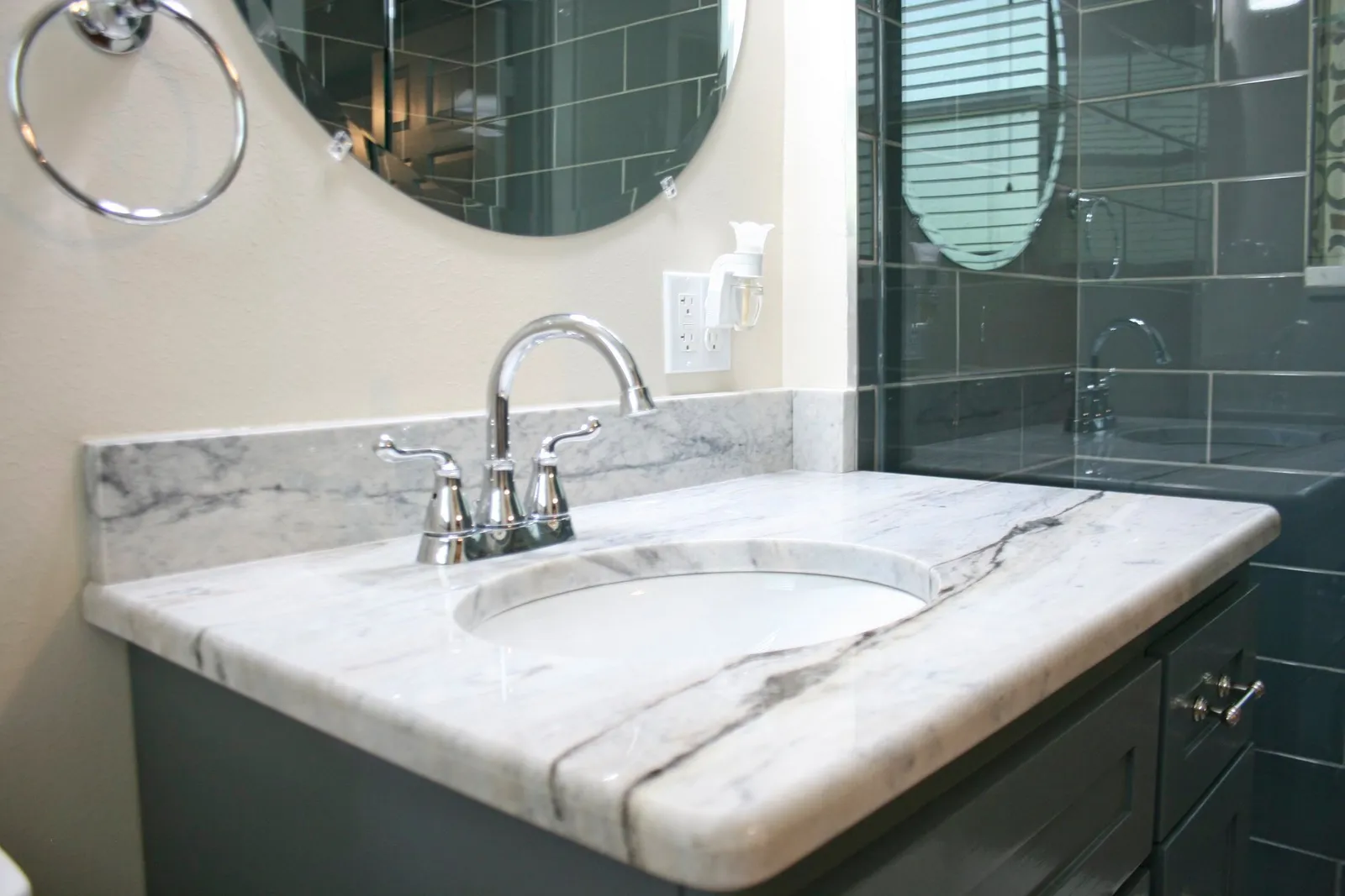 Marble vanity top on a gray cabinet with a round mirror and dark green wall tile.