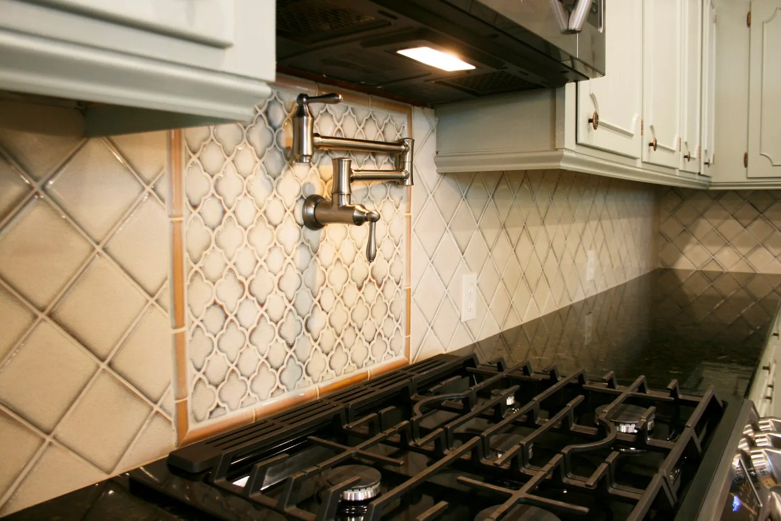 Close-up of a diamond-pattern tile backsplash behind a gas cooktop and pot filler.