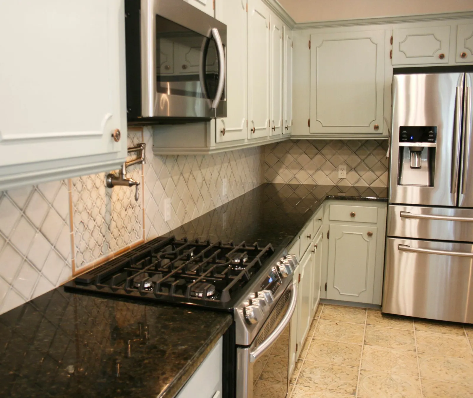 Gas range and stainless appliances between sage cabinets, with a diamond-pattern tile backsplash.