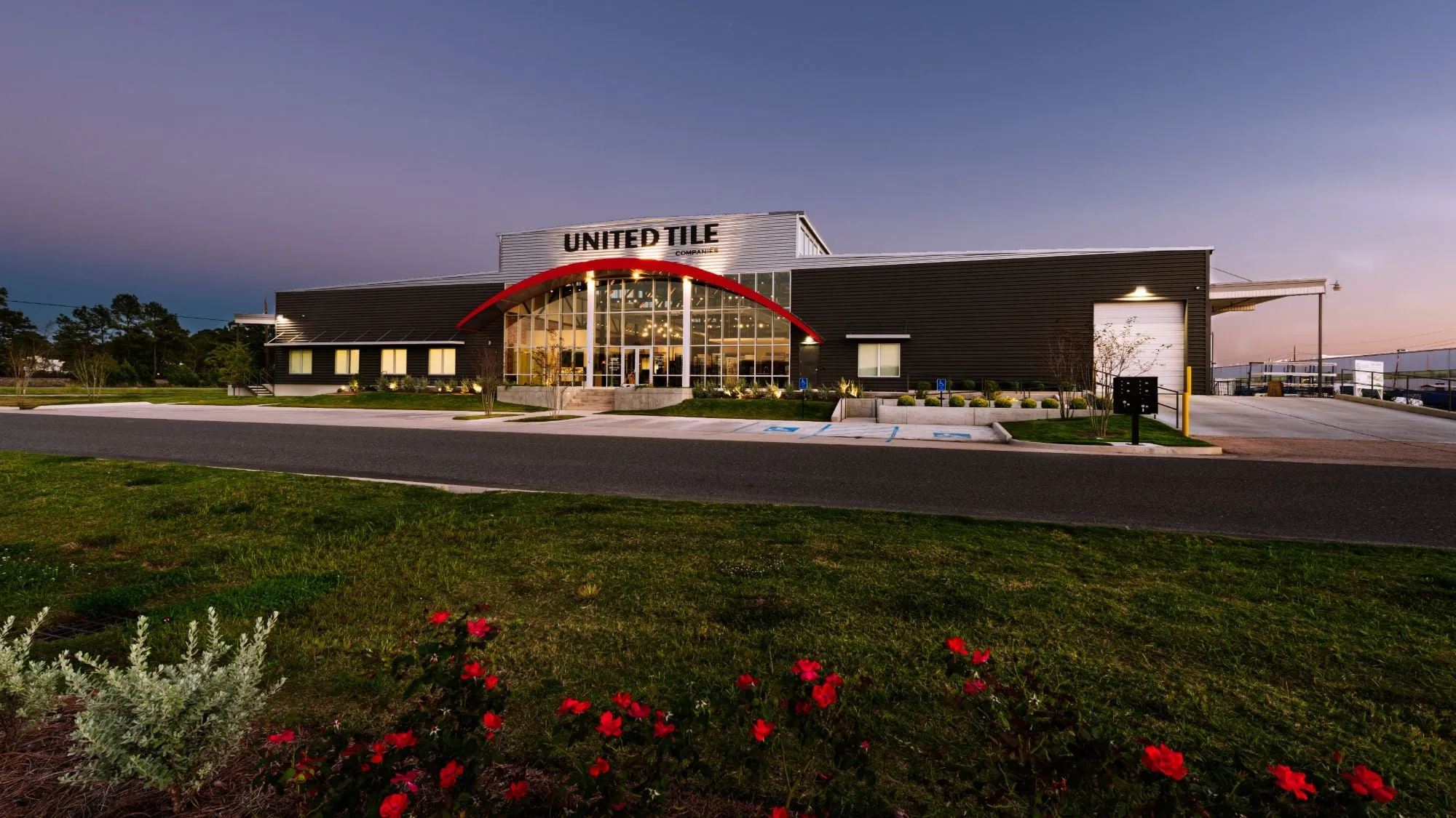 The United Tile building at dusk, its red arch and glass entrance lit from within, with red roses blooming in the lawn across the street.