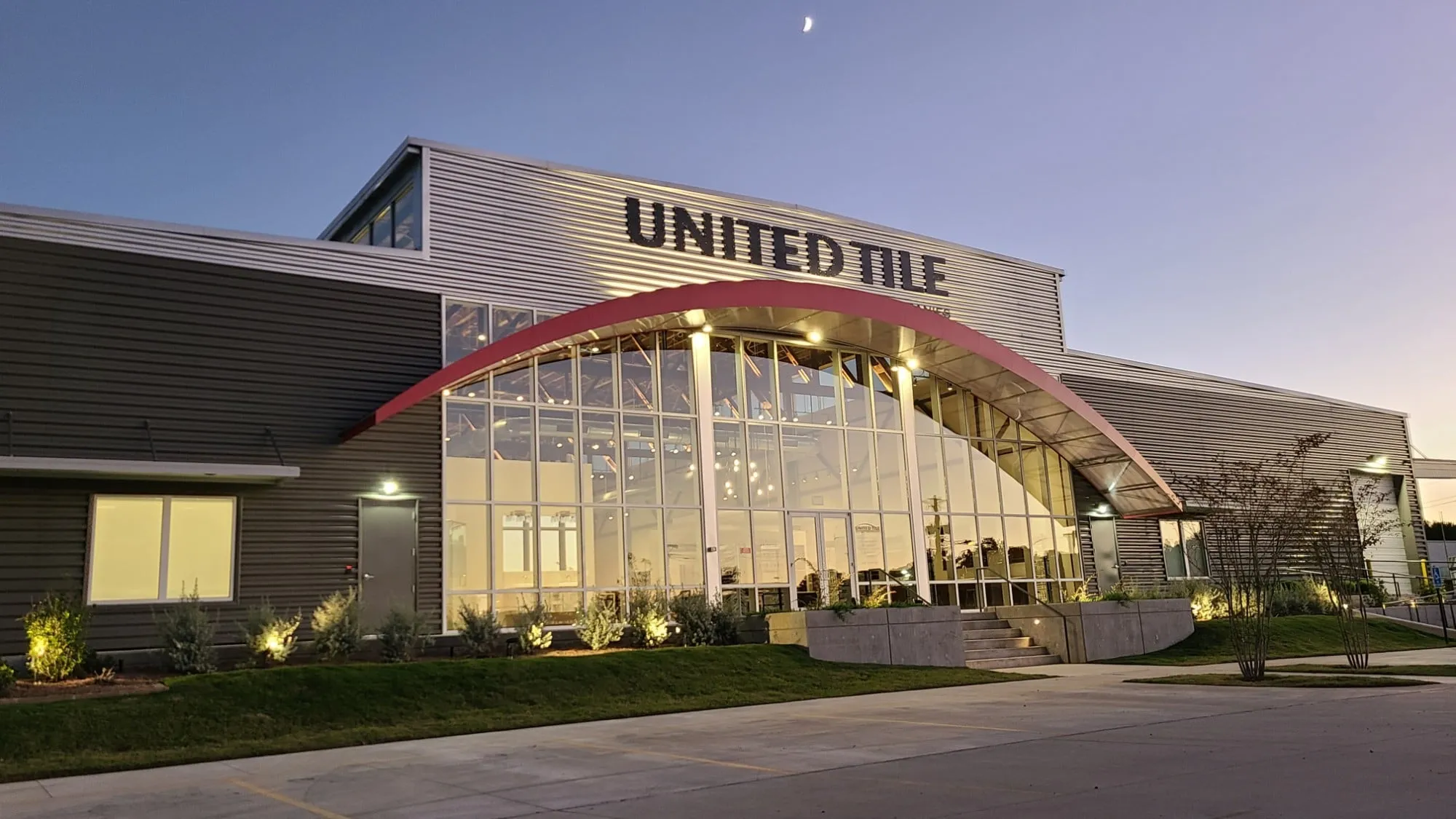 The United Tile showroom at dusk: the red arch over the tall glass entrance glows from inside, below the United Tile sign.