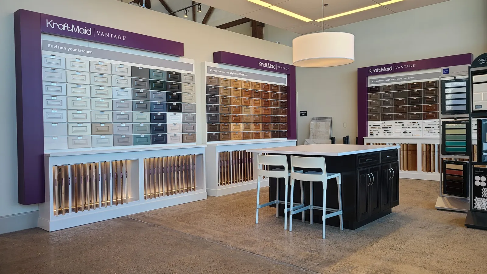Our cabinet displays: walls of KraftMaid Vantage door samples in painted colors and wood stains, a hardware wall, and a dark cabinet island with two stools.
