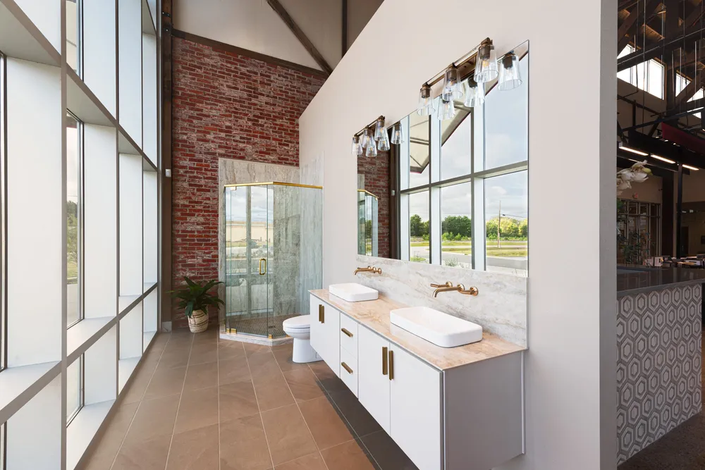 A bathroom display in our showroom: a gold-framed glass shower enclosure against old brick, and a double vanity under two wide mirrors.
